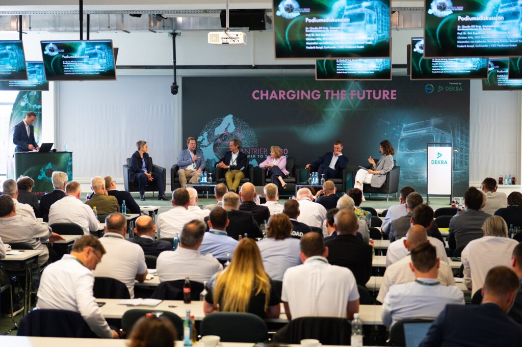Konferenzbühne mit Panel von sechs Personen in Sesselreihen, Hintergrundbanner „CHARGING THE FUTURE“ und moderner Grafik; Moderator am Pult links, überwiegend Geschäftsleute im Publikum.