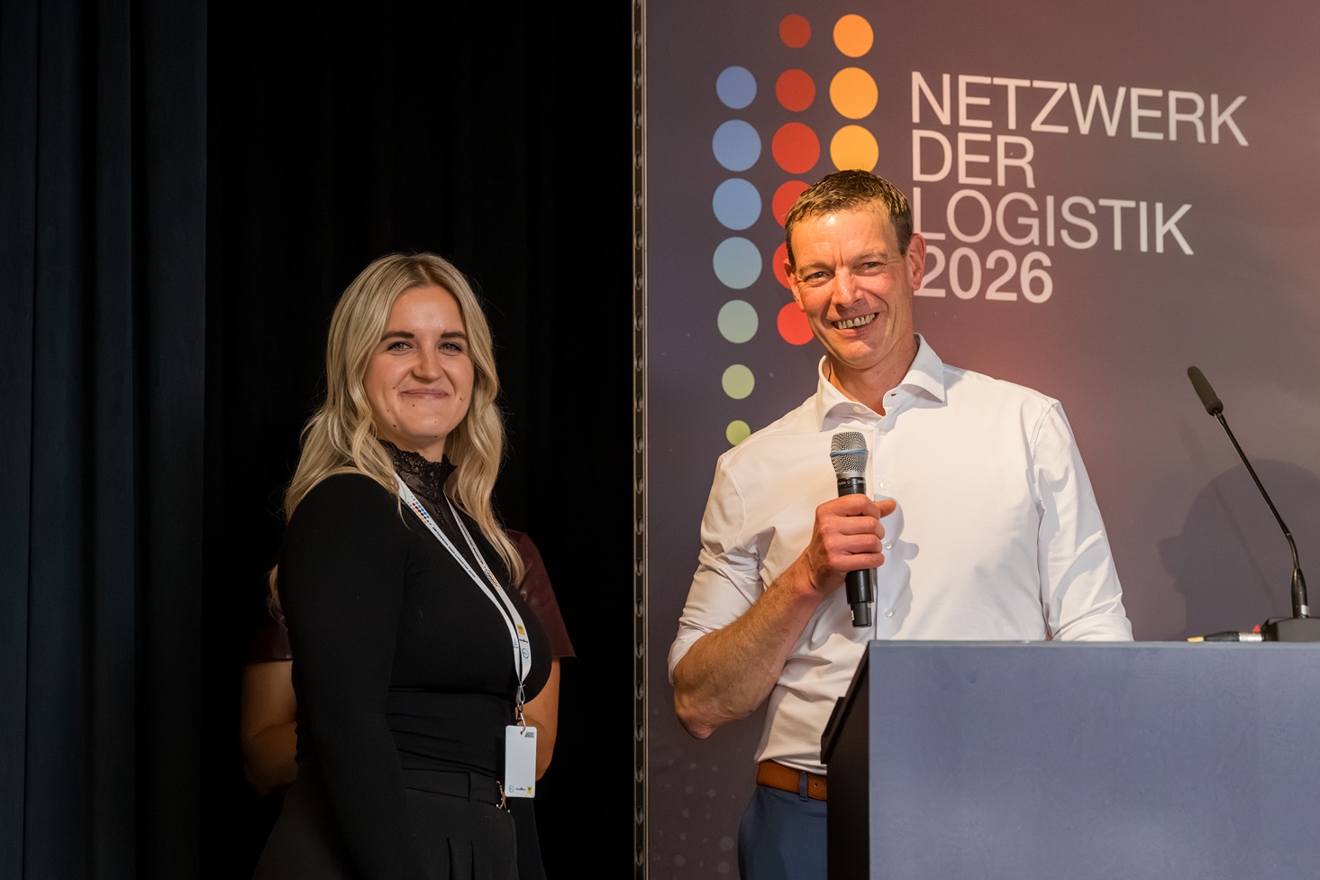 Zwei Personen auf Bühne bei Netzwerk der Logistik 2026; Frau links lächelnd, Mann rechts mit Mikrofon am Rednerpult.
